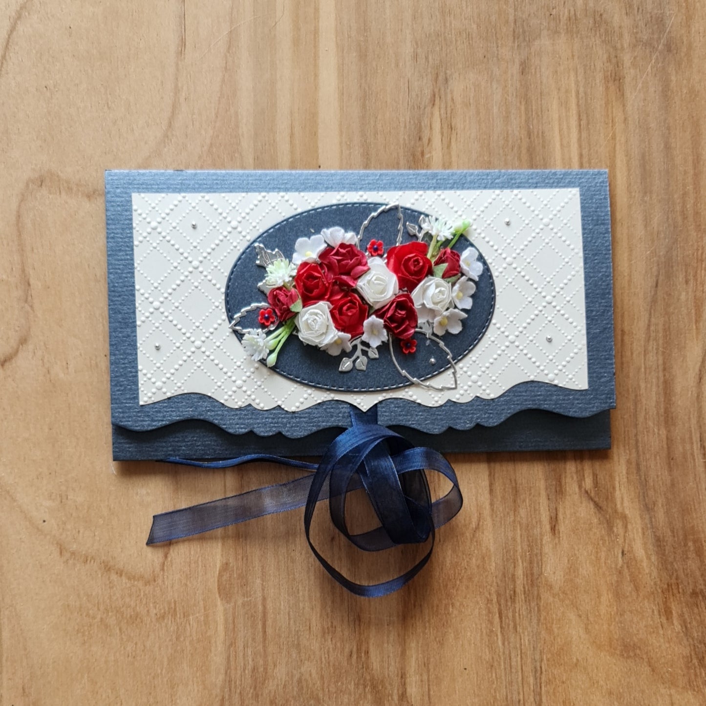 Dāvanu aploksne perlamutra zilā krāsā ar 3D sarkaniem un kremkrāsas ziediem uz gaiša fona un tumši zilu lentas aizdari 10,8 x 19 cm (APU2) - www.tiptip.lv 